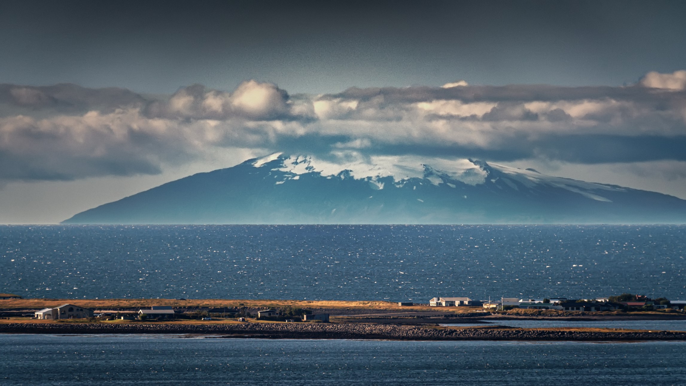 landscape photography showing a tall mountain glacier in the distance, with water in the foreground and some small houses on a protruding bit of land in the middle of the image