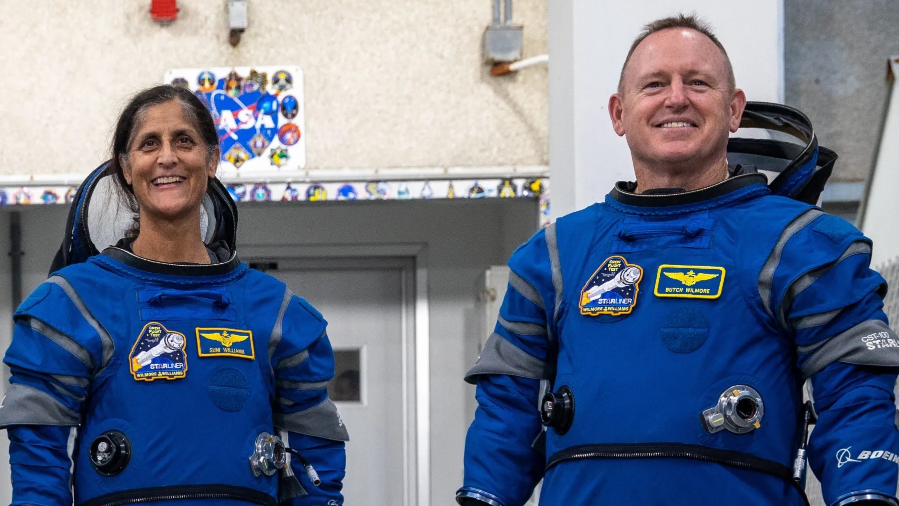 two astronauts in spacesuits smiling and standing in front of a wall. the wall has a nasa logo on it surrounded by mission patches