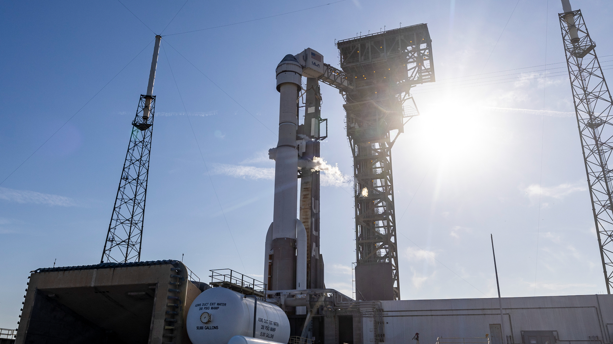 a rocket on a launch pad with the sun behind it and a blue sky, surrounding by towers