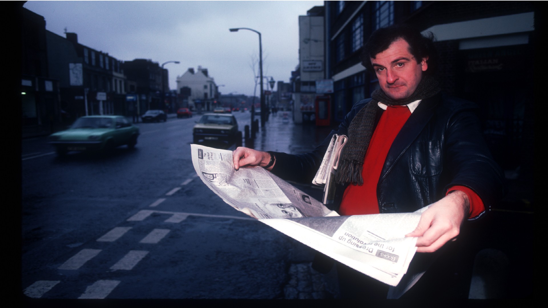 a man in a black jacket sits outside reading a newspaper